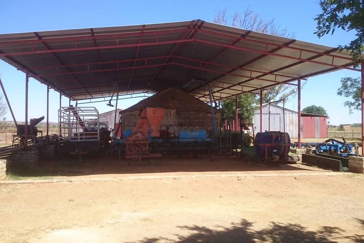 A large open-sided farm utility shed with a metal roof and steel support structure, providing covered storage for agricultural equipment.