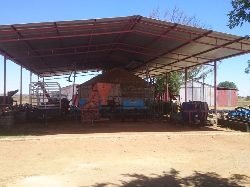 A large open-sided farm utility shed with a metal roof and steel support structure, providing covered storage for agricultural equipment.
