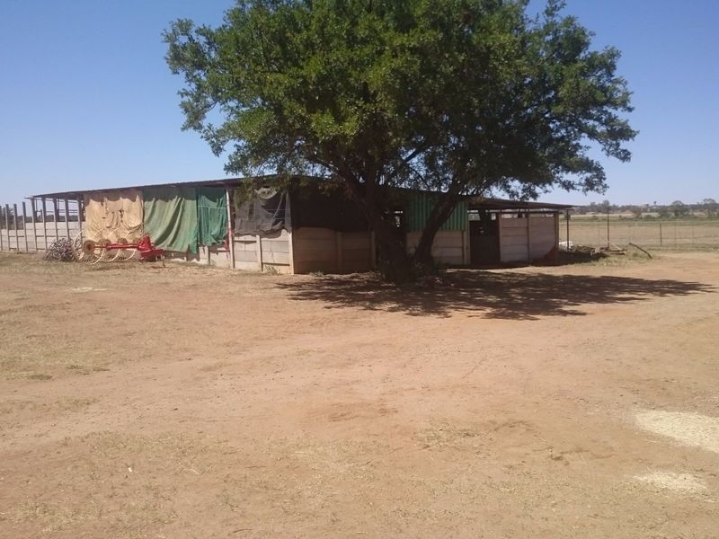 An outdoor farm utility structure featuring concrete walling and a metal roof, situated in a large open yard area.