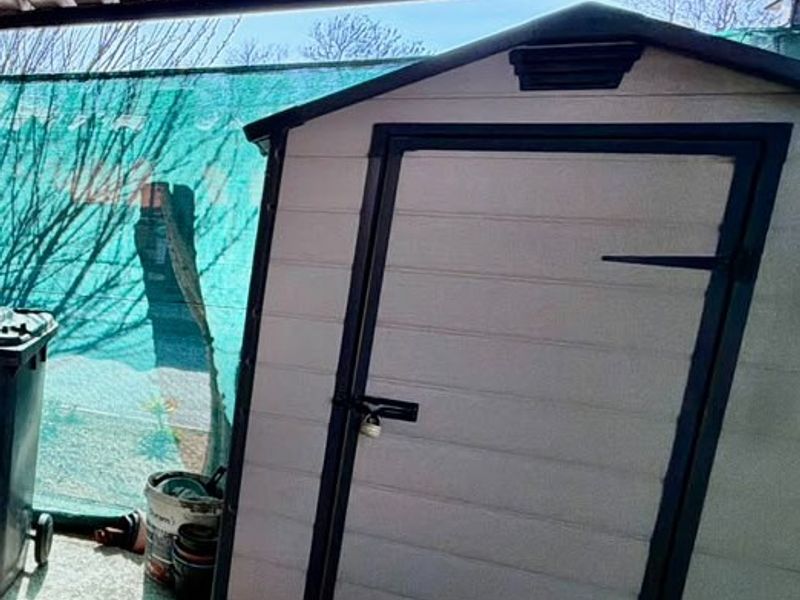 A plastic storage shed situated under a corrugated metal roof structure in an outdoor utility area.