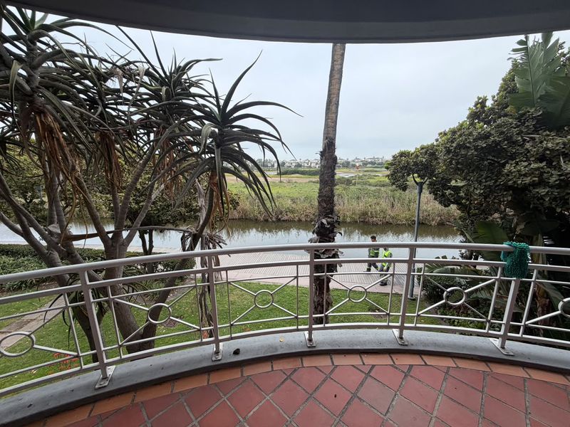 A curved balcony featuring a metal balustrade and terracotta tiled flooring, offering scenic views of the surrounding garden, water feature, and landscape.