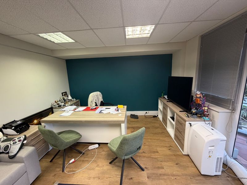 A functional office space featuring laminate flooring, a suspended ceiling with recessed lighting, and a bold teal accent wall with large window access.