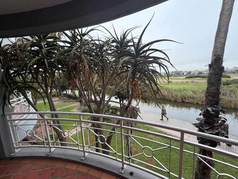 A covered balcony featuring terracotta tiling and a metal balustrade, offering scenic views of the adjacent garden, waterway, and walking path.