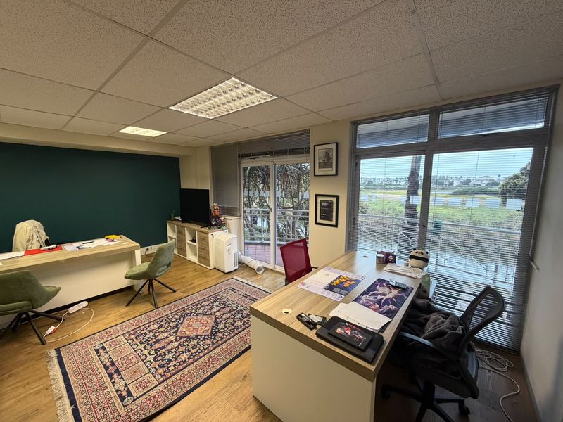 A bright office space featuring laminate flooring, suspended ceilings with recessed lighting, and large windows providing natural light and exterior views.