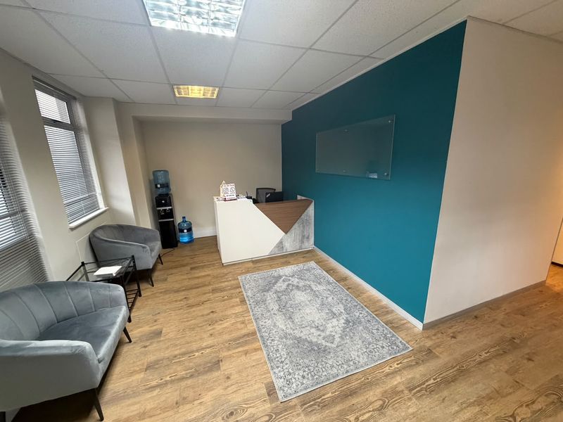 A professional office reception area featuring wood-look laminate flooring, a modern reception desk, and a bold teal accent wall with a glass display board.