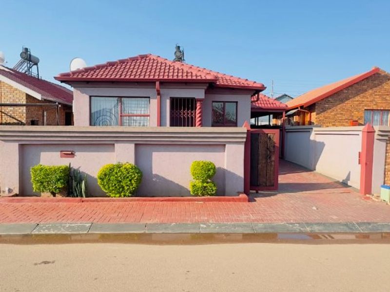 Front view of a single-story house featuring a tiled roof, perimeter wall, and a paved driveway with a secure entrance gate.