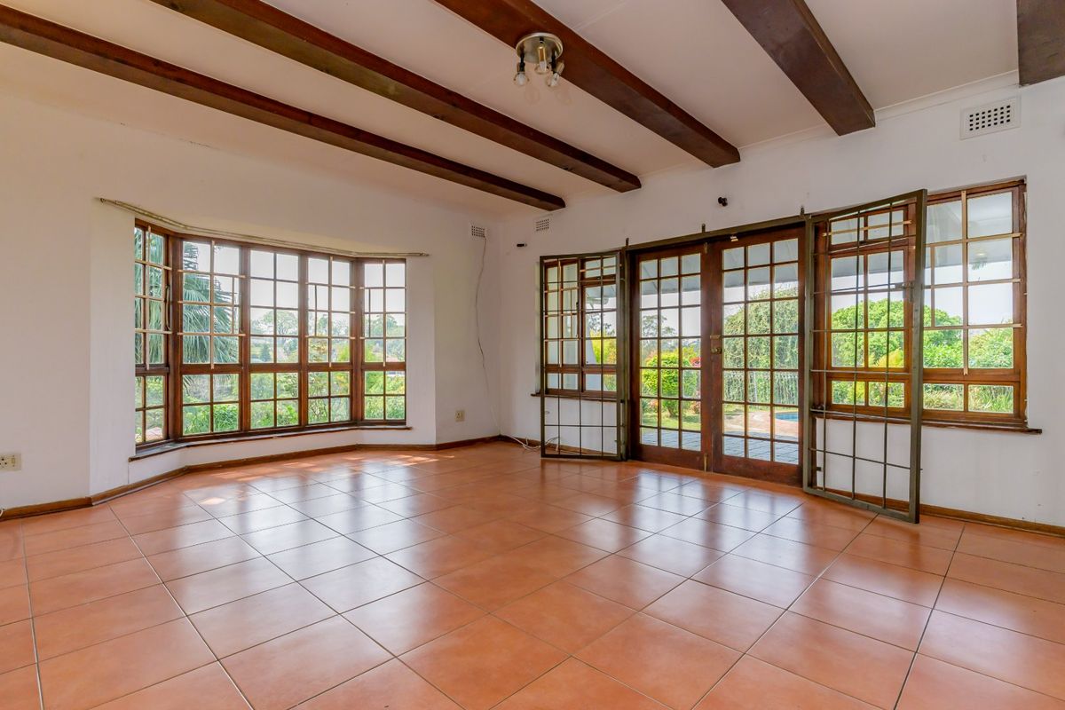 Spacious living area featuring exposed timber ceiling beams, large bay windows, and tiled flooring with direct access to the garden through timber-framed glass doors.