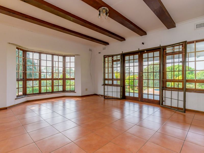 Spacious living area featuring exposed timber ceiling beams, large bay windows, and tiled flooring with direct access to the garden through timber-framed glass doors.