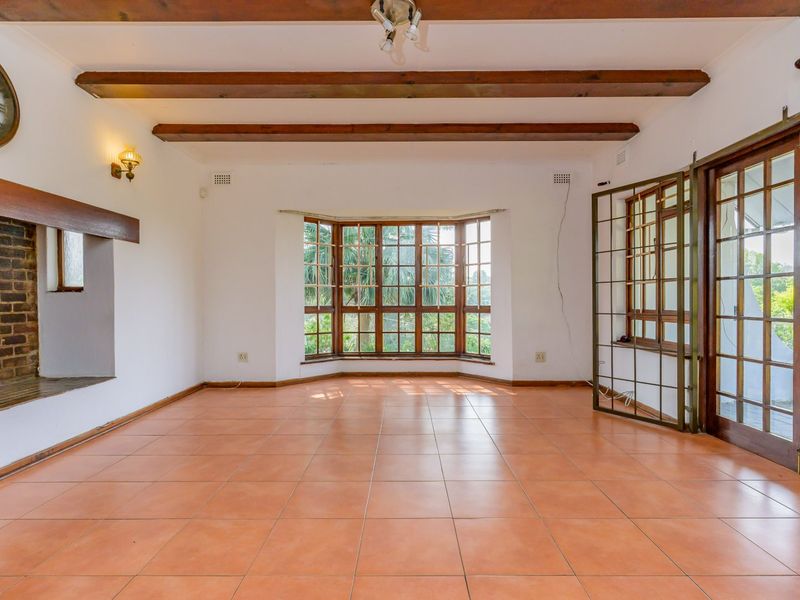 Spacious living room featuring exposed timber ceiling beams, tiled flooring, a brick fireplace, and a large bay window with access to the exterior.