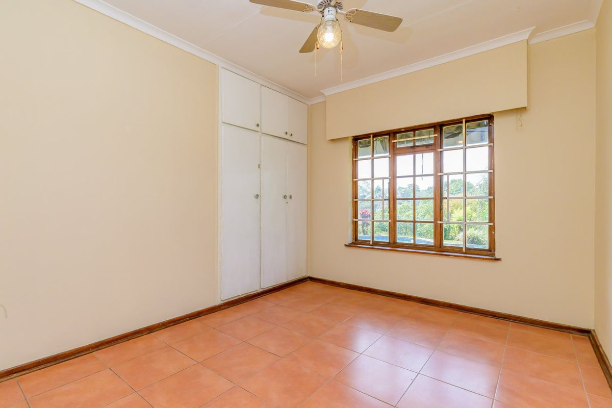 An empty bedroom featuring tiled flooring, built-in cupboards, a ceiling fan, and large wooden-framed windows overlooking the garden.