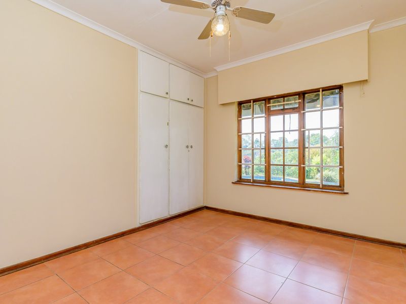 An empty bedroom featuring tiled flooring, built-in cupboards, a ceiling fan, and large wooden-framed windows overlooking the garden.