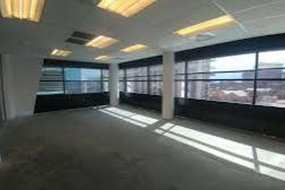 An empty, open-plan commercial office space featuring large windows, structural columns, and a suspended ceiling with integrated lighting.
