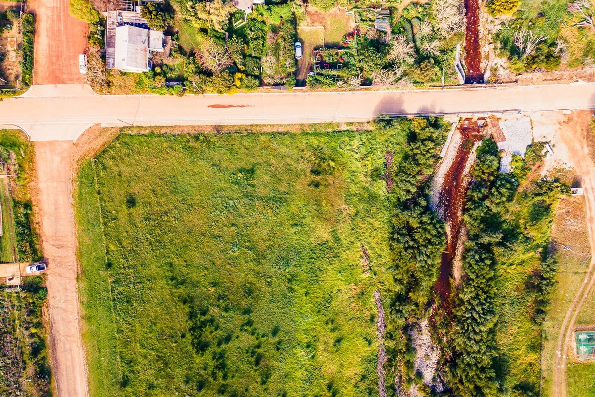Aerial view of a vacant residential plot featuring natural vegetation, a stream, and direct access to a dirt road.