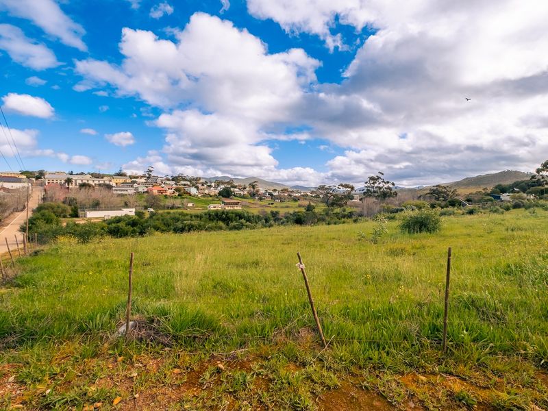 A wide-angle view of a vacant residential plot featuring natural grass terrain and wire fencing, overlooking a distant hillside neighborhood.