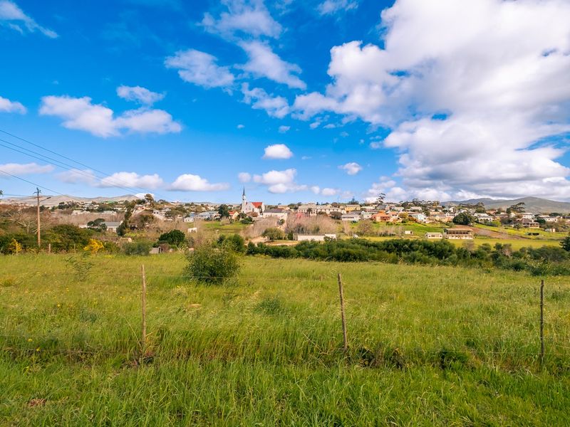 A scenic view of a rural landscape featuring open grassy fields, wire fencing, and a distant town with a church steeple under a blue sky.