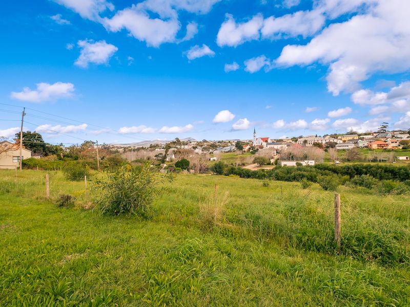 A scenic view of a vacant residential plot featuring grassy terrain, wire fencing, and a distant view of the surrounding town and landscape.