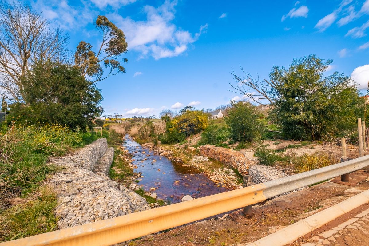 A scenic view of a natural stream featuring stone gabion retaining walls and surrounding vegetation, viewed from a roadside guardrail.
