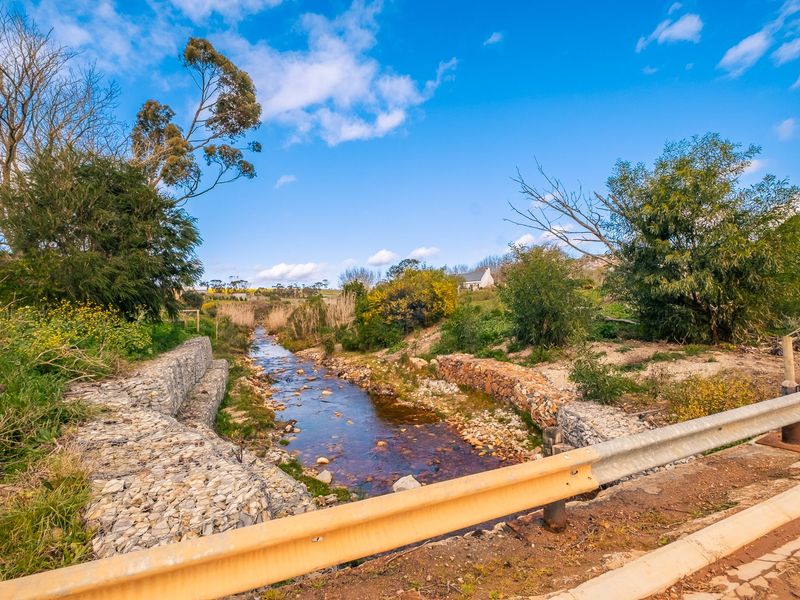 A scenic view of a natural stream featuring stone gabion retaining walls and surrounding vegetation, viewed from a roadside guardrail.