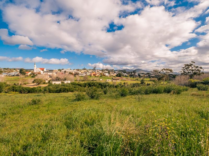 A scenic view of a grassy vacant residential plot overlooking a distant town and rolling hills under a bright blue sky.