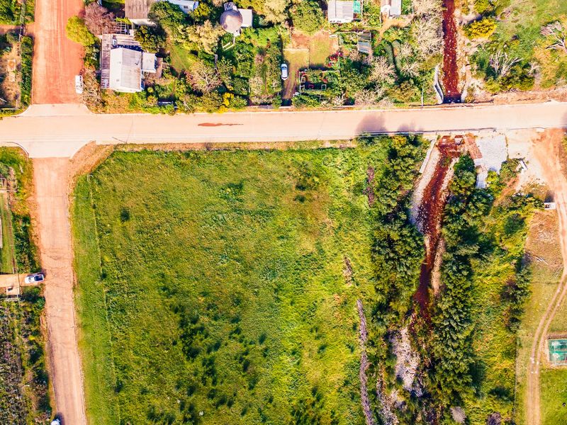 Aerial view of a vacant residential plot featuring natural vegetation, a stream, and direct access to a dirt road.
