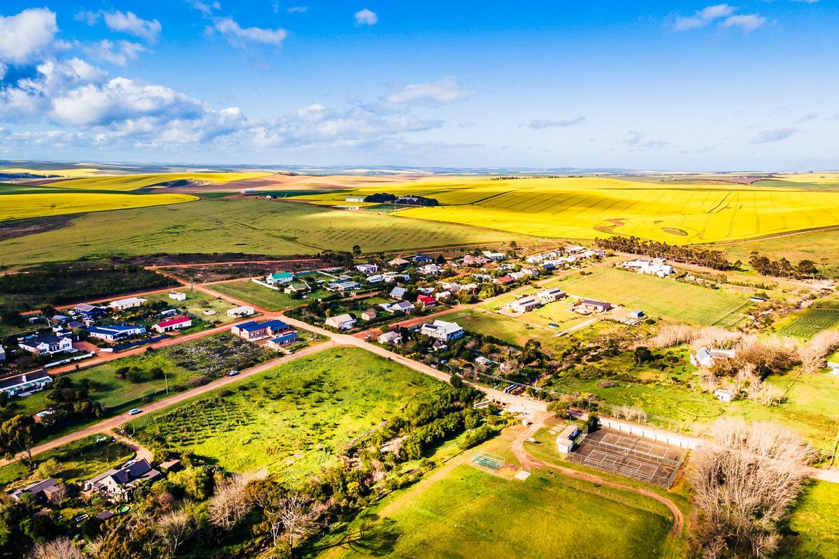 An aerial view of a rural residential area featuring open fields, established housing plots, and a tennis court facility.