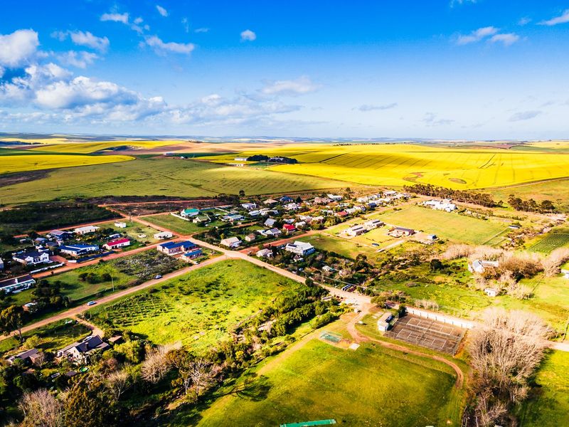 An aerial view of a rural residential area featuring open fields, established housing plots, and a tennis court facility.