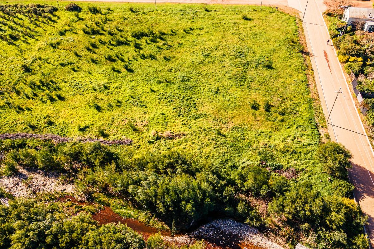 An aerial view of a vacant residential plot featuring natural grassland, road access, and a bordering creek.