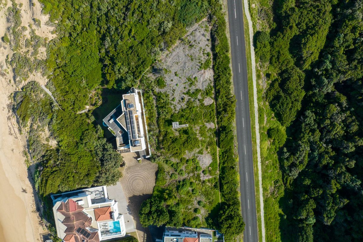 Aerial view of a vacant residential plot situated between a coastal area and a main road, surrounded by established vegetation.