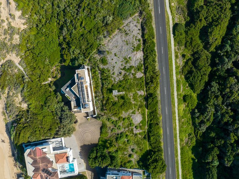 Aerial view of a vacant residential plot situated between a coastal area and a main road, surrounded by established vegetation.