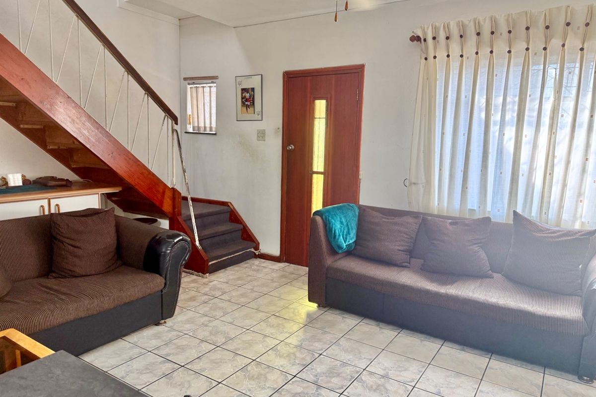 A living room featuring tiled flooring, a timber staircase, and a ceiling fan, offering a functional layout with potential for cosmetic updates.