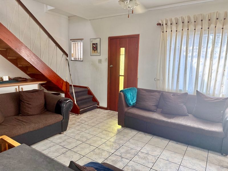 A living room featuring tiled flooring, a timber staircase, and a ceiling fan, offering a functional layout with potential for cosmetic updates.