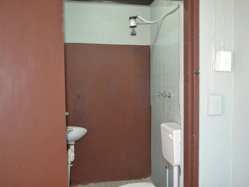 A basic, dated bathroom featuring a wall-mounted basin, shower head, and toilet cistern with exposed plumbing and simple tiled finishes.