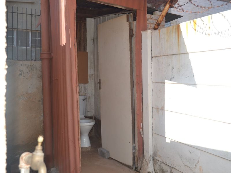 An outdoor utility toilet structure featuring a basic toilet bowl, housed within a corrugated metal enclosure adjacent to a precast concrete boundary wall.
