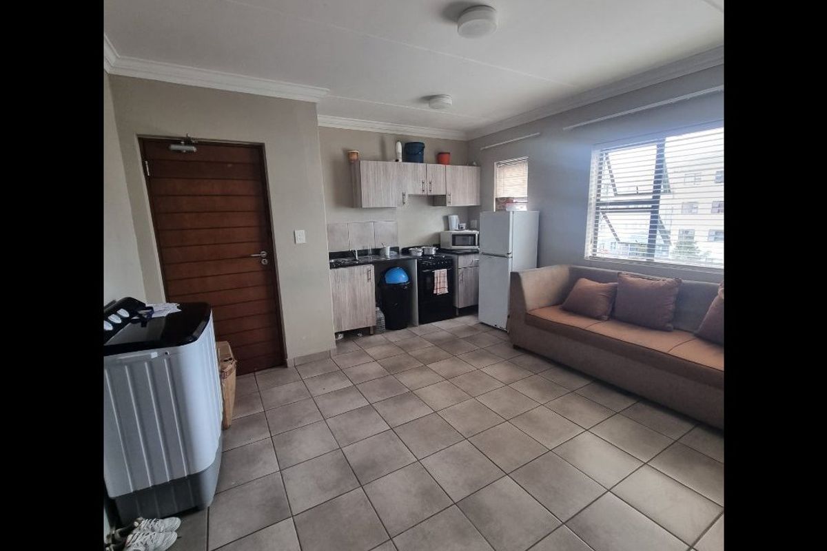 Open-plan living area featuring tiled flooring, a compact kitchen with built-in cabinetry, and large windows with venetian blinds.