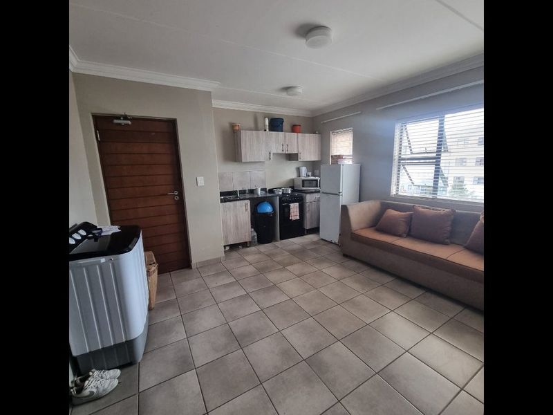 Open-plan living area featuring tiled flooring, a compact kitchen with built-in cabinetry, and large windows with venetian blinds.