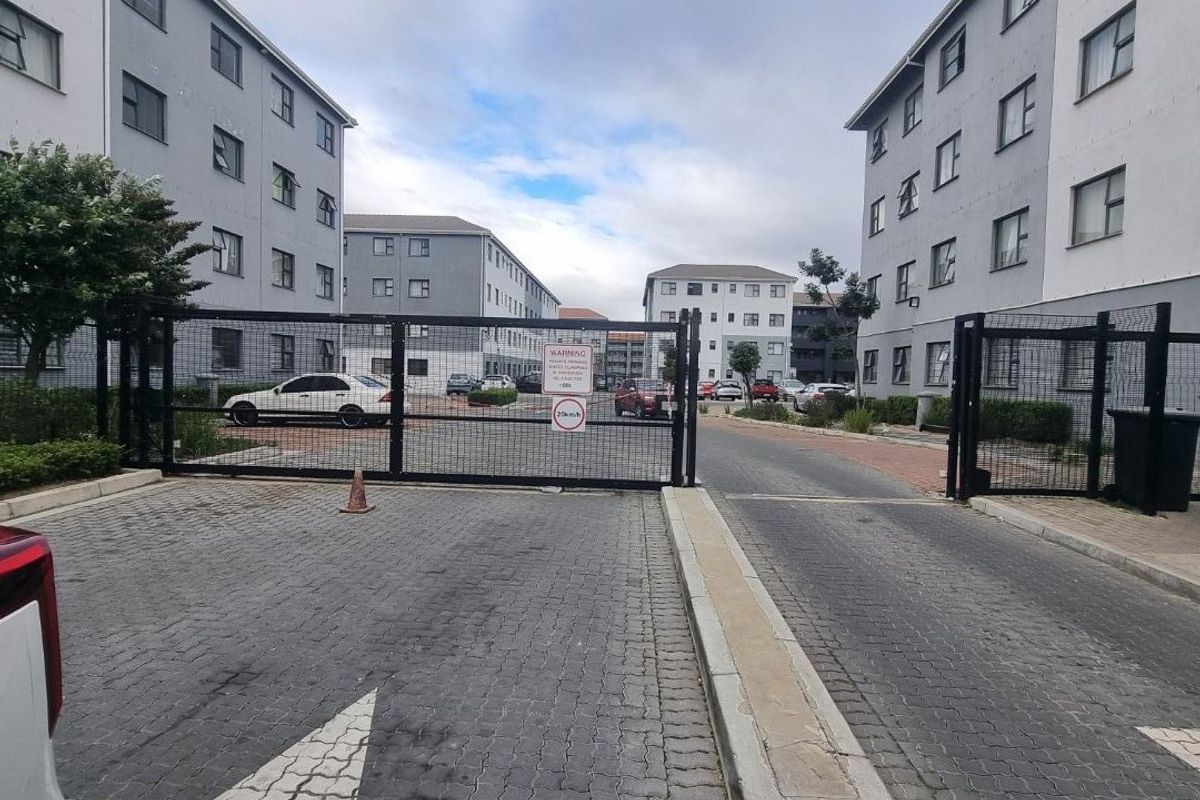 View of the apartment complex entrance featuring a secure gated access point, paved driveway, and multi-story residential buildings.
