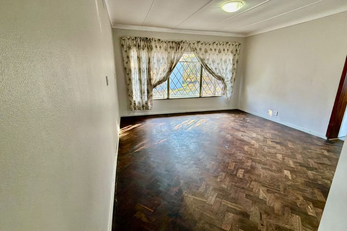A spacious, empty living room featuring classic parquet flooring, security burglar bars on the window, and neutral wall finishes.