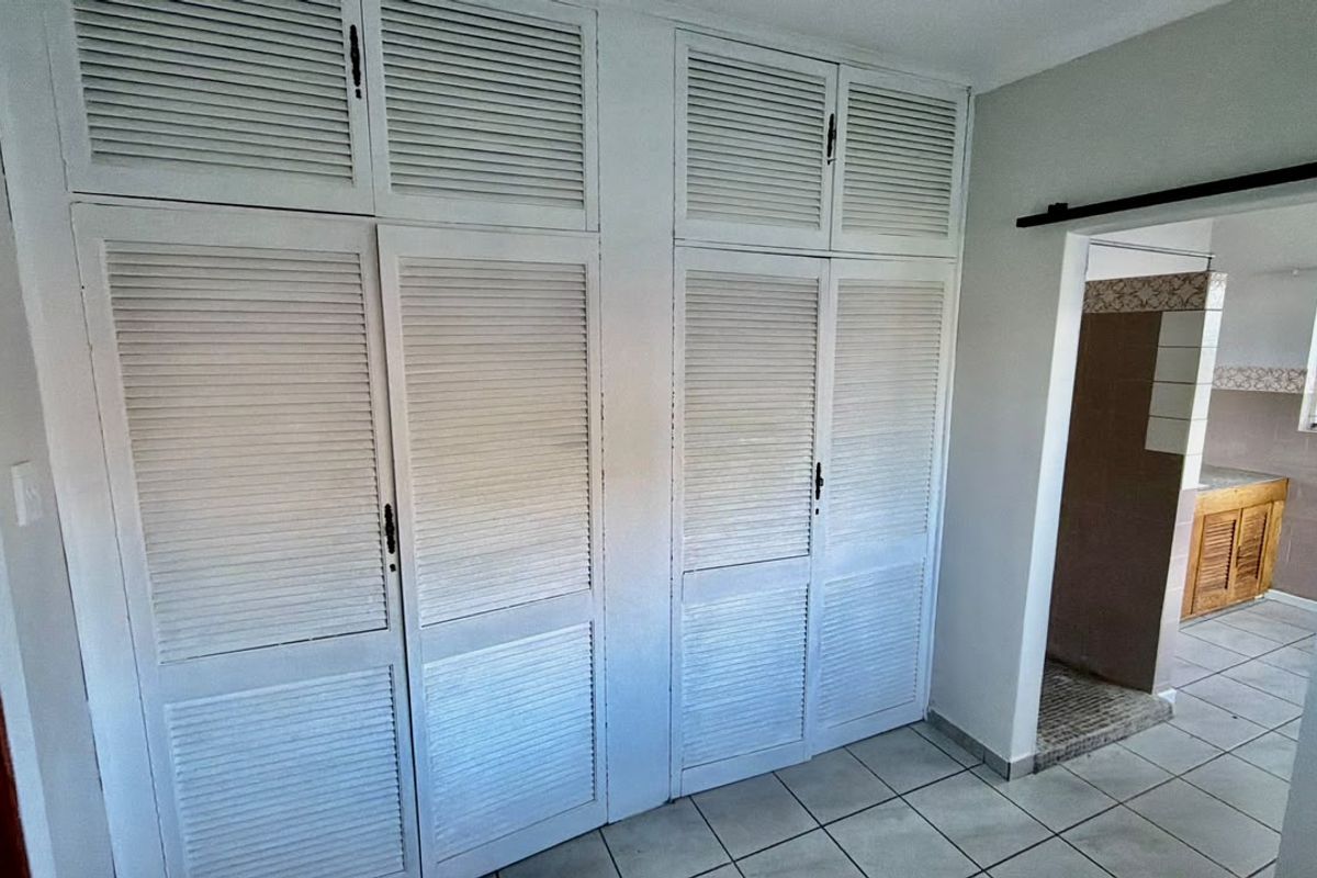 A bedroom featuring white louvered built-in cupboards and tiled flooring, with a view into an adjacent tiled bathroom area.