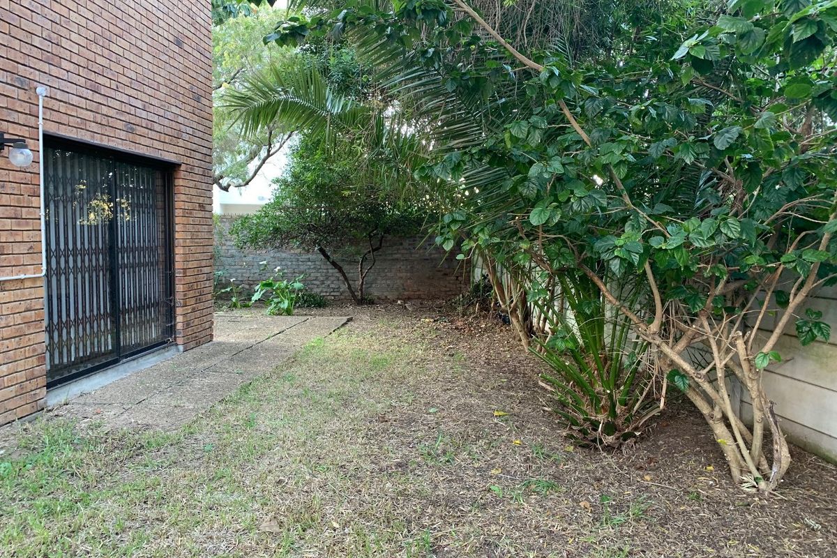 A rear exterior view of the townhouse featuring a brick facade, security gate, and a small garden area with established trees and boundary walls.