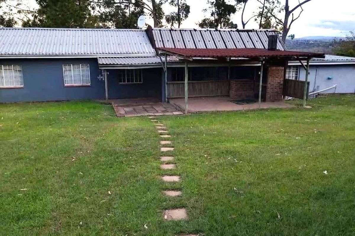 Rear view of the property featuring a covered patio with a built-in brick braai, corrugated metal roofing, and a stone pathway leading across the lawn.