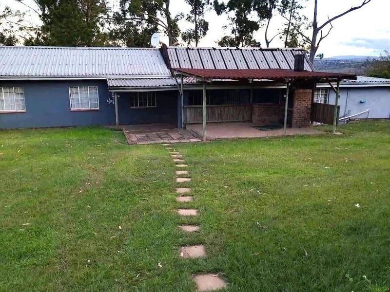 Rear view of the property featuring a covered patio with a built-in brick braai, corrugated metal roofing, and a stone pathway leading across the lawn.