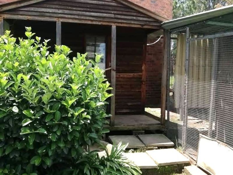 A rustic timber-clad structure with a covered porch, situated adjacent to a large brick silo and a wire mesh enclosure in a garden setting.