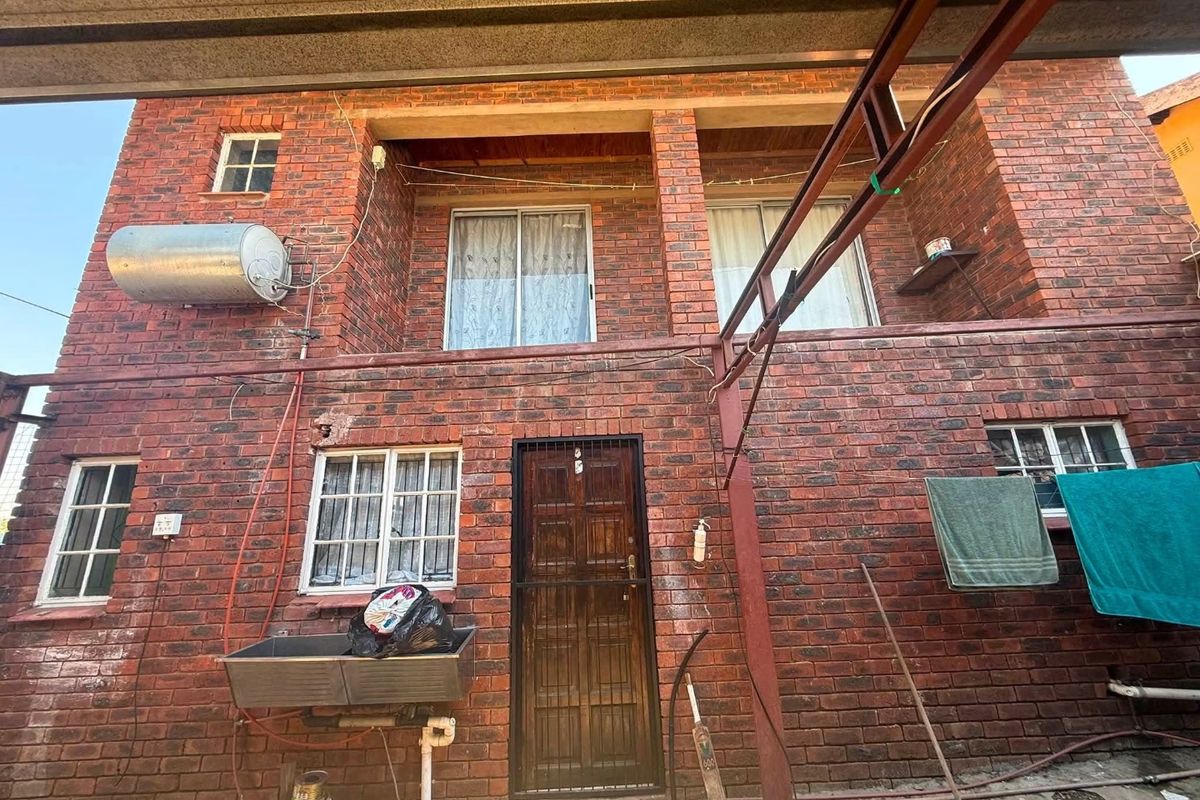 Rear exterior view of a double-story face-brick house featuring an external geyser, laundry sink, and security gate access.