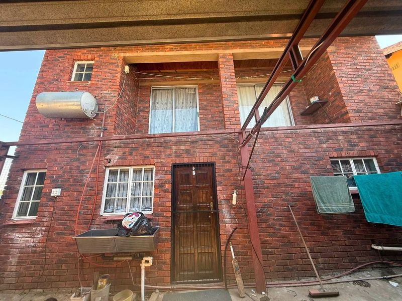 Rear exterior view of a double-story face-brick house featuring an external geyser, laundry sink, and security gate access.