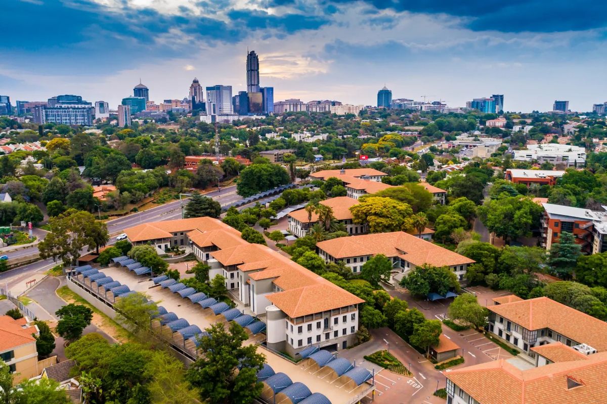 Aerial view of a commercial office park complex featuring terracotta tiled roofs, extensive landscaped grounds, and covered parking facilities with city skyline backdrop.