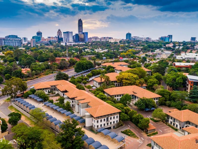 Aerial view of a commercial office park complex featuring terracotta tiled roofs, extensive landscaped grounds, and covered parking facilities with city skyline backdrop.