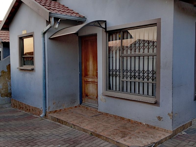 Front elevation of a single-story house featuring a tiled porch, security gate, and paved driveway, showing signs of exterior wear and plaster damage.