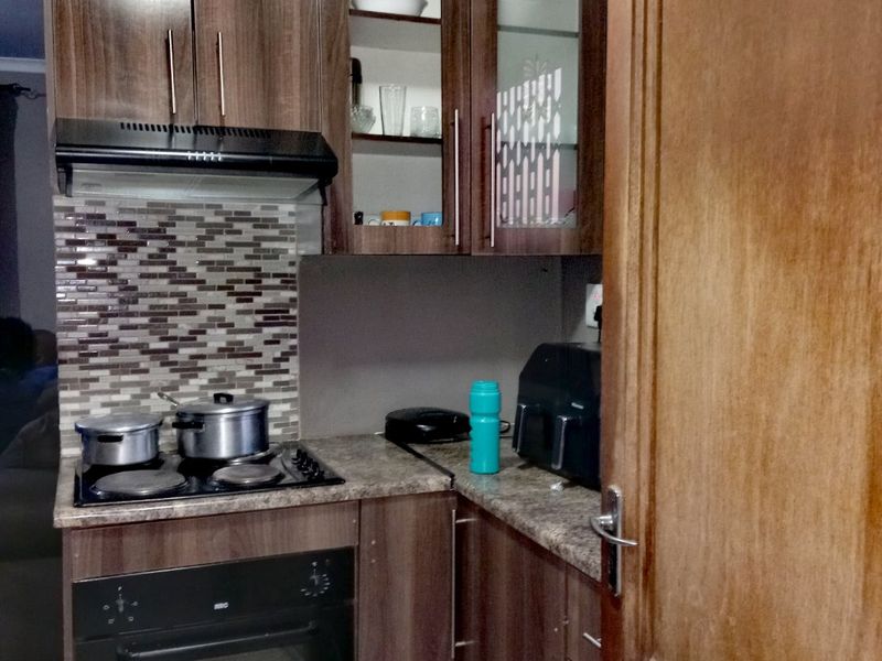 Compact kitchen featuring wood-look laminate cabinetry, mosaic tile backsplash, built-in oven, and electric hob with an integrated extractor fan.