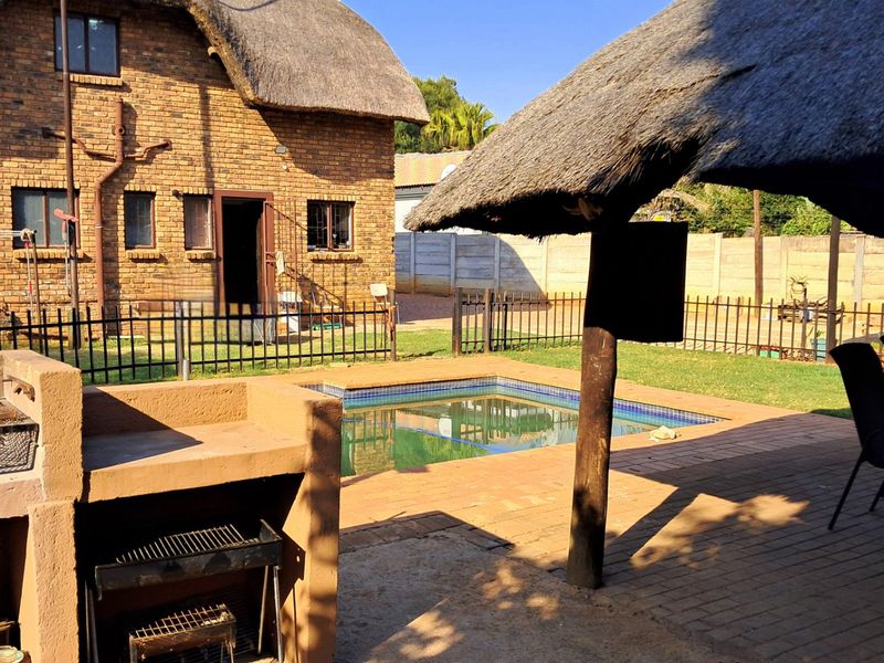 Outdoor entertainment area featuring a swimming pool, paved patio, and a built-in braai, set against a traditional thatched-roof brick house.