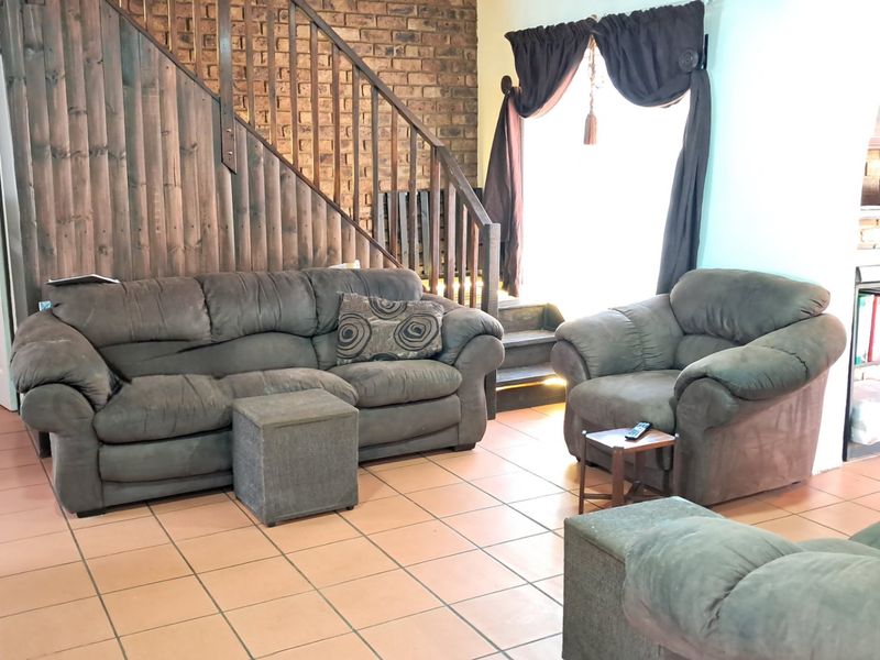 Spacious living room featuring a rustic brick accent wall, timber staircase with balustrade, and durable tiled flooring throughout.